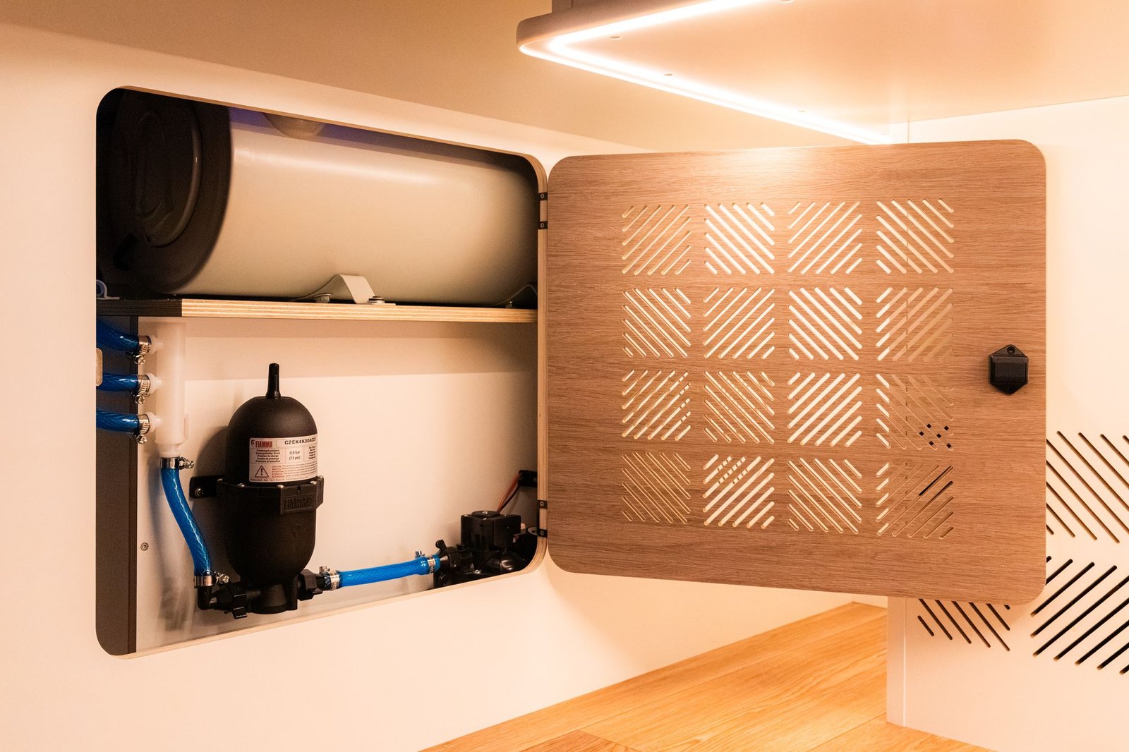 Sistema de agua bajo el mueble: depósito, bomba y tuberías tras una puerta de madera calada