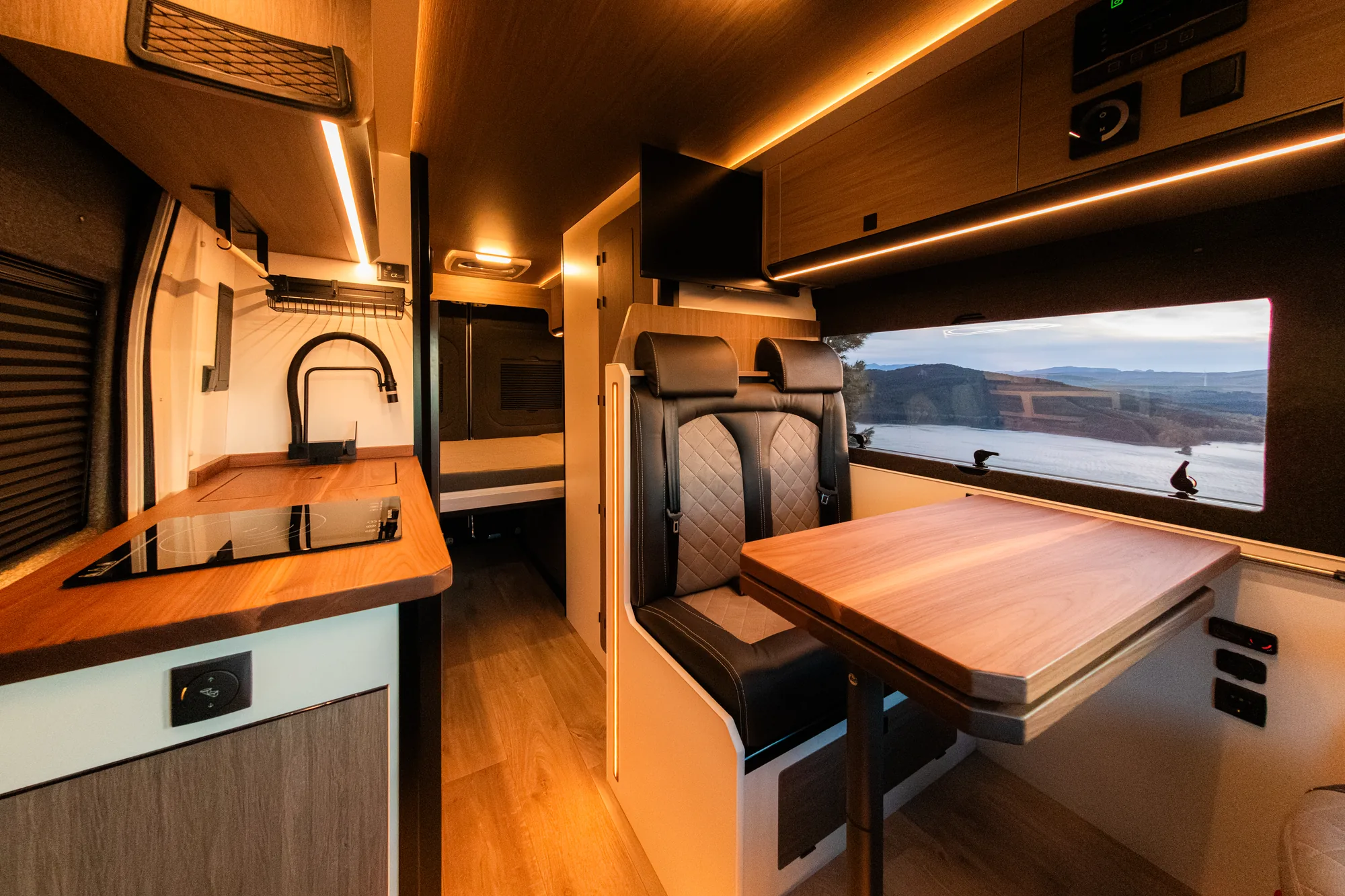 Interior camperizado con cocina de inducción, fregadero y comedor con mesa de madera junto a una ventana con vistas al embalse